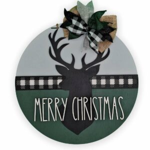 A round Christmas decoration featuring a black silhouette of a merry Christmas deer in the center. "Merry Christmas" is written below the reindeer. The top boasts a plaid and burlap bow with green leaves, while a black and white plaid strip adorns the middle, making it an ideal custom door sign.