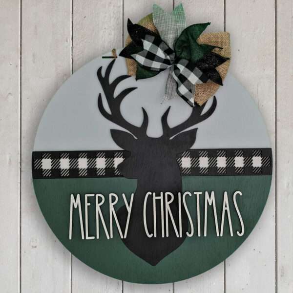 A round Christmas decoration featuring a black silhouette of a merry Christmas deer in the center. "Merry Christmas" is written below the reindeer. The top boasts a plaid and burlap bow with green leaves, while a black and white plaid strip adorns the middle, making it an ideal custom door sign. The sign is placed on a white wood background.