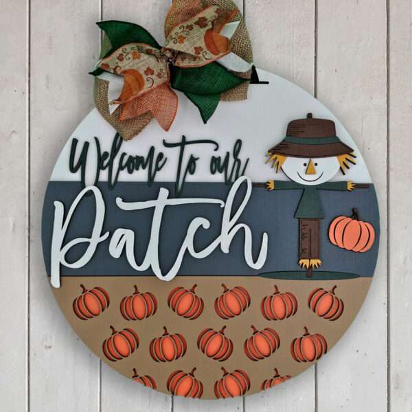 A round "welcome to our patch" door sign on a wooden background features a crafty scarecrow, pumpkins, and the text "Welcome to our Patch" in white letters. The sign is decorated with a colorful burlap ribbon at the top.