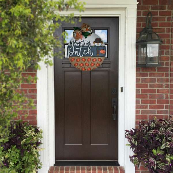 A dark brown front door decorated with a round fall-themed wreath featuring the text "Happy Fall Y'all" and small pumpkins. The brick wall house façade has a lantern light fixture to the right, potted plants on both sides of the door, and a charming "Welcome to Our Patch" door sign.