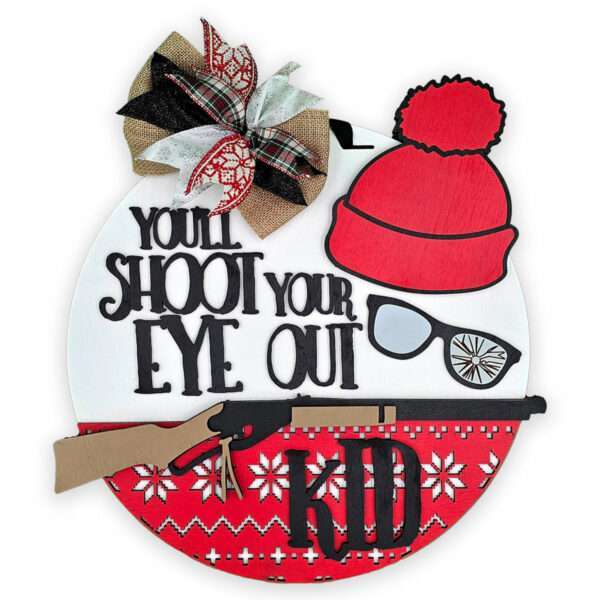 A round holiday decoration features a plaid bow, a red knit hat, glasses, and a rifle. The text reads, "You'll shoot your eye out Kid" over a red and white snowflake pattern at the bottom, referencing a famous quote from the classic Christmas Story movie.