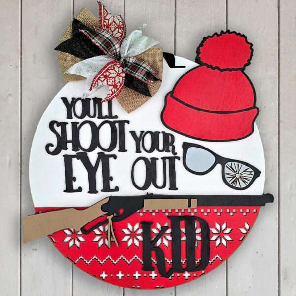 A round wall decoration against a wooden background features a holiday theme with a red and white knit pattern. It has a red pom-pom hat, broken glasses, and a toy rifle with the text "You'll shoot your eye out, kid." A large bow sits at the top left, reminiscent of the Christmas Story movie.