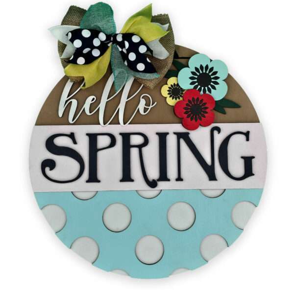 A round, decorative wooden sign reads "hello SPRING" in large, playful black letters. The sign is painted in pastel colors and features polka dots on the lower half. A colorful bow and spring poppies adorn the top, adding a vibrant touch to the cheerful design.