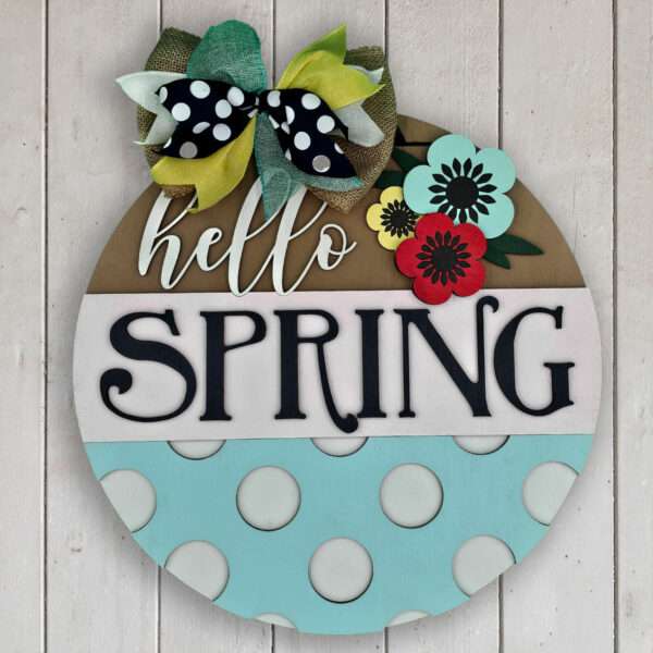 A circular wooden sign against a white background reads "hello SPRING" in black letters. It is decorated with a multicolored bow, flowers, and vibrant spring poppies. The bottom section features large white polka dots on a light blue background.