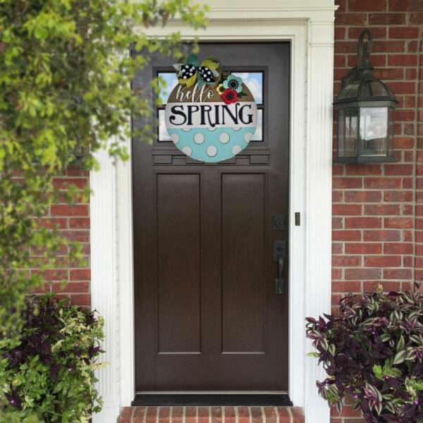 A brown front door with an illuminated custom "Hello Spring" sign featuring blue polka dots hangs in the center. The door is framed by white trim and brick walls. There is a lantern-style light fixture to the right, potted plants and vibrant spring poppies on both sides of the door.