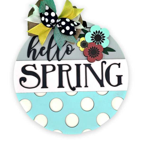 A round wooden sign features "hello SPRING" in black letters. It's decorated with a large, multicolored bow and poppies in teal, red, and yellow. The bottom section displays a light teal background with large white polka dots.