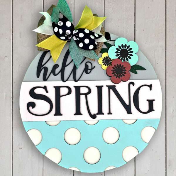 A decorative round wooden sign hangs on a wall, featuring the words "hello SPRING" in stylish black fonts. The top has multicolored flowers, including vibrant Spring Poppies, and a bow with black and white polka dots and yellow ribbon. The bottom half of the sign is a pale blue with white polka dots.