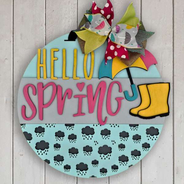 A round sign reads "Hello Spring" with "Hello" in yellow and "Spring" in pink. It features an umbrella, rain boots, and a decorative bow. The bottom section has a pattern of clouds with raindrops. The background is a white wooden surface, perfectly capturing the essence of spring showers.