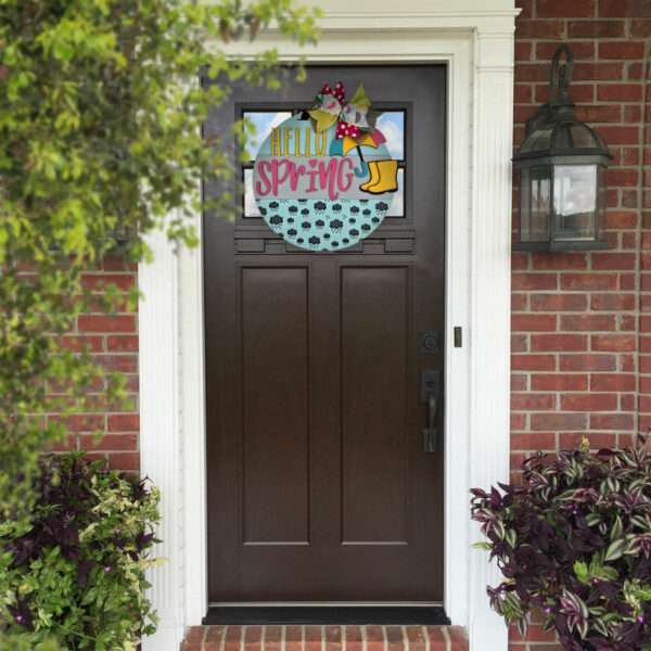 A dark brown front door with a spring-themed wreath and custom door sign hanging on it. The wreath features a yellow birdhouse, colorful flowers, and the words "Hello Spring" in multicolored letters. The door is framed by red brick and white trim, with rain boots filled with plants and a wall-mounted light fixture nearby.