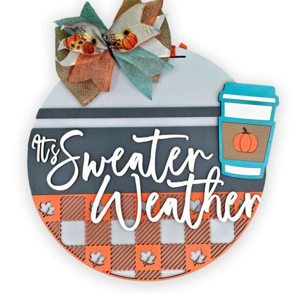 A custom door sign reads "It's Sweater Weather" in white cursive on a round background with striped patterns and small leaf cutouts. A bow with orange accents is at the top, and a coffee cup with a pumpkin design graces the right side.