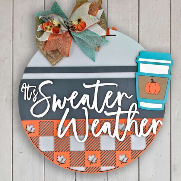 A round wooden sign with a fall theme displays the phrase "It's Sweater Weather" in white cursive. It features a colorful fabric bow adorned with pumpkins at the top and a small coffee cup illustration on the right, showcasing an orange pumpkin on the cup. The background has checkered and striped patterns with small white leaves.