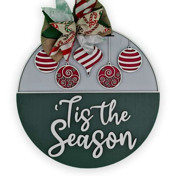 Round holiday decoration featuring a festive bow, red and white ornaments, and the text "'Tis the Season" in white. This custom door sign has the top half adorned with ornaments, while the bottom half displays the cheerful text.