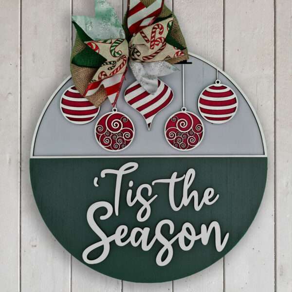 Round holiday decoration featuring a festive bow, red and white ornaments, and the text "'Tis the Season" in white. This custom door sign has the top half adorned with ornaments, while the bottom half displays the cheerful text. The sign is displayed on a white wood background.