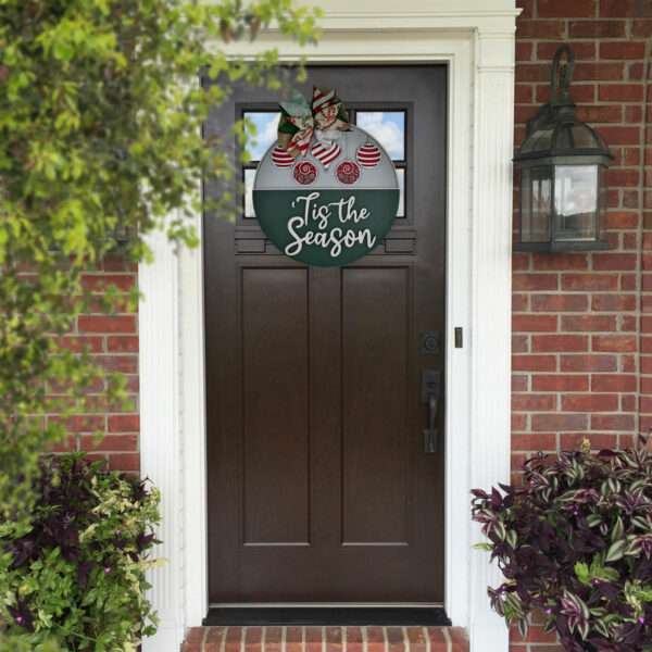 Round holiday decoration featuring a festive bow, red and white ornaments, and the text "'Tis the Season" in white. This custom door sign has the top half adorned with ornaments, while the bottom half displays the cheerful text. The sign is displayed on the front door of a house with a dark door.