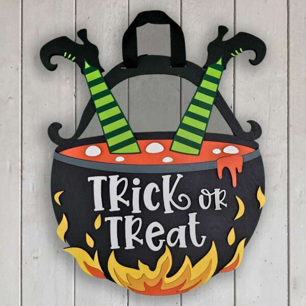 A Halloween decoration features a bubbling cauldron with "Trick or Treat" written on its front. Green striped witch legs with curly-toed shoes stick out from the top, while yellow flames surround the base. The background is a rustic wooden wall.