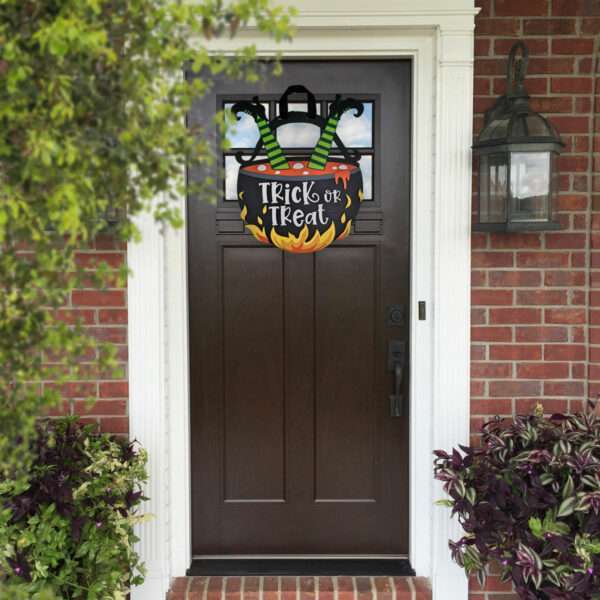 A brown front door is decorated with a sign that reads "Trick or Treat" in white letters, perfectly capturing the Halloween spirit. The sign features a witch's legs sticking out of a cauldron. The red brick exterior of the house is partially visible, along with some potted plants near the entrance.