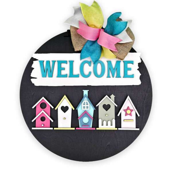 A round black sign with the word "WELCOME" in blue letters on a white strip at the top. Below are colorful, decorative birdhouses, adding charm to the design. The sign is topped with a bow made of pink, yellow, blue, and burlap ribbons.