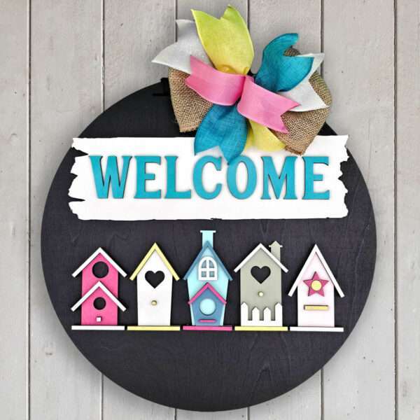 Round wooden welcome sign with "WELCOME" in turquoise letters on a white banner. Below the banner, five colorful birdhouses are lined up. A bow made of burlap and brightly colored ribbons adorns the top of the sign. The background is a white wooden panel.