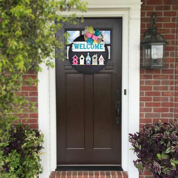 A brown front door with white trim is adorned with a colorful custom "WELCOME" sign featuring birdhouses. The door is framed by red brick walls and flanked by a lantern-style light fixture and lush green plants.