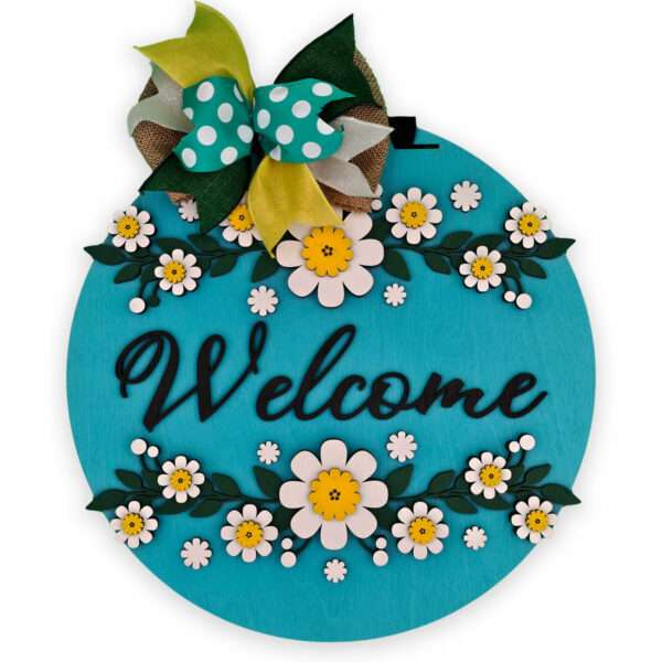 A circular, turquoise "Welcome" sign adorned with white and yellow daisies and green leaves along the top and bottom edges. At the top, there's a decorative bow with green, yellow, and white polka-dotted teal ribbons.