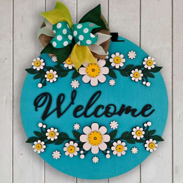 A round, turquoise wooden sign with the word "Welcome" written in cursive. The sign is decorated with white and yellow daisies, green leaves, and a large bow in green, yellow, polka-dotted white, and turquoise at the top. The sign hangs on a white wooden background.