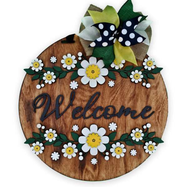 A round, wooden door sign with the word "Welcome" in black cursive letters. The sign is decorated with white and yellow daisies and green leaves. A bow with black and white polka dots, green, and burlap ribbons is affixed at the top.