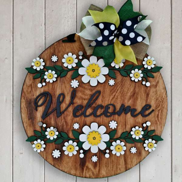 A round wooden welcome sign adorned with white and yellow flowers, including charming daisies, and green leaves. A yellow, green, and black polka-dotted bow sits at the top. The background is a light wooden plank wall.