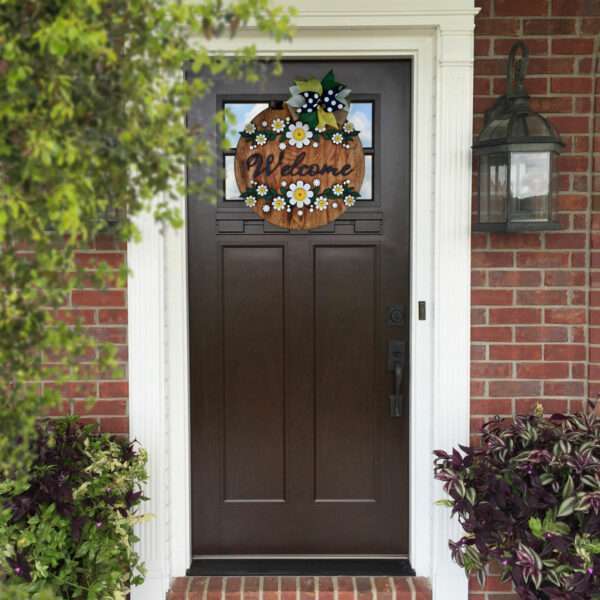 A dark brown front door with a white frame, featuring a decorative wreath that says "Welcome." The wreath is adorned with white daisies and blue flowers, along with a black-and-white checkered bow. Surrounding the entrance are green plants and a brick exterior wall, completing this welcoming scene.