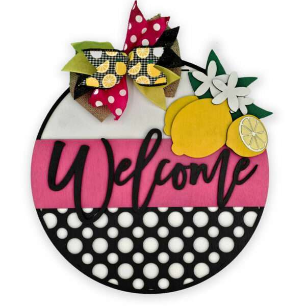 A circular welcome sign decorated with a pink stripe across the middle, black and white polka dots on the bottom, and a colorful bow with lemon patterns at the top. The sign features lemon and flower cutouts along with the word "Welcome" in cursive script.