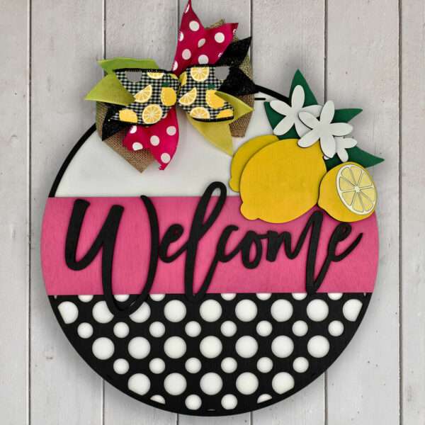 A circular wooden "Welcome" sign adorned with a bold black script on a pink, white, and black polka-dotted background. The top features a decorative bow with various patterns and colors, alongside wooden lemon and flower embellishments. The sign hangs on a light wooden wall.