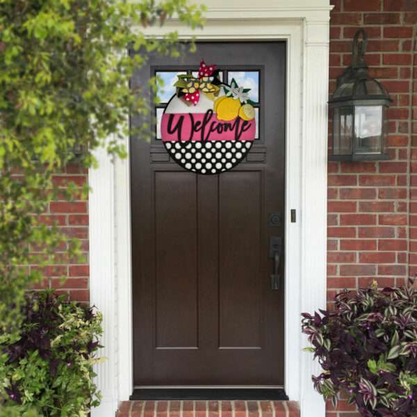 Front door of a brick house with a dark brown panel door. The door features a custom "Welcome" sign with lemons, flowers, and polka dots hanging in the glass panel at the top. To the right, there's a vintage-style outdoor lantern light.