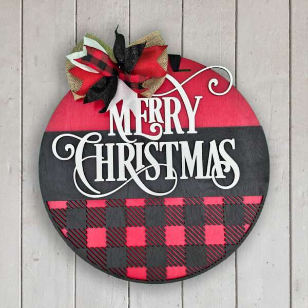 A round Christmas decoration with the words "Merry Christmas" in white letters on a red and black plaid background. The top of the decoration has a festive bow with red, green, and black ribbons. The decoration is hung on a white wooden surface.