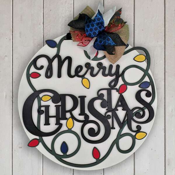 A round wooden Christmas ornament with the words "Merry Christmas" written in script. Colorful lightbulb designs surround the text, and a decorative bow made of black, blue, gold, and red ribbon is attached at the top. The background is white wooden planks.