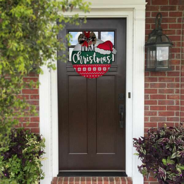 A dark wooden front door is adorned with a festive wreath featuring the words "Merry Christmas" in white letters on a red and green background. The door is surrounded by a brick exterior, with potted plants and a rustic lantern on either side.