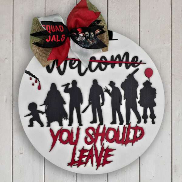 A round Halloween sign against a white wooden background. The sign features the silhouettes of horror characters with blood-red text saying "WELCOME YOU SHOULD LEAVE" over a crossed-out "Welcome." It is decorated with a bow at the top.