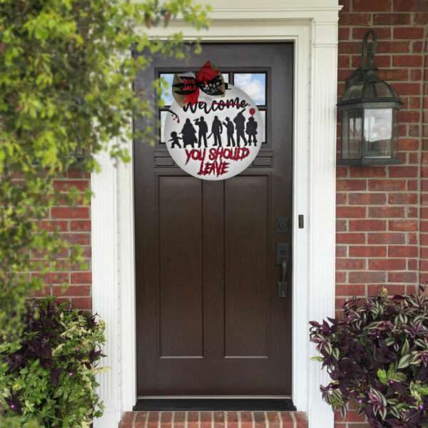 A brown front door with a custom door sign featuring a circular Halloween theme hangs from the top. The sign showcases silhouettes of spooky figures such as zombies and a witch, with the text "Welcome...You Should Leave" in red and black. The door is framed by red brick walls.