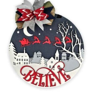 A circular Christmas door sign featuring a nightscape with Santa in his sleigh pulled by reindeer flying over a snowy village and tree silhouettes. The word "BELIEVE" is scripted in red at the bottom. A bow in red, green, and white adorns the top.