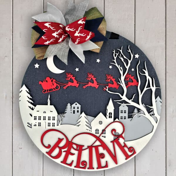 A Christmas-themed circular sign with a winter village nightscape and Santa's sleigh with reindeer flying across the night sky. The word "BELIEVE" is prominently cut out in red at the bottom. A festive bow adorns the top.