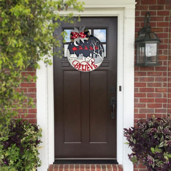 A brown front door adorned with a Christmas-themed wreath featuring Santa Claus and reindeer flying over a winter village scene, with the word "BELIEVE" in bold red letters. The door is set in a charming brick house, surrounded by a lantern sconce and lush plants.