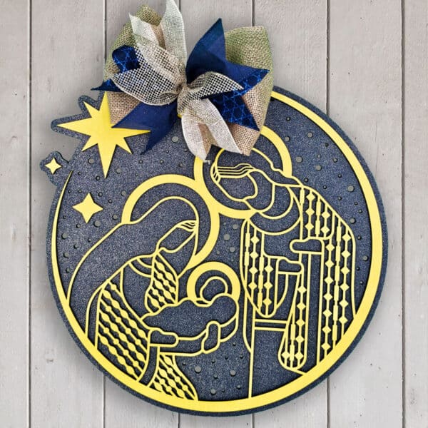 A circular Christmas nativity decoration with stylized, laser-cut figures of Mary, Joseph, and baby Jesus in yellow against a blue background. The top is adorned with a large bow featuring blue, white, and gold ribbons and can be used as a festive door sign. The design includes a star motif.