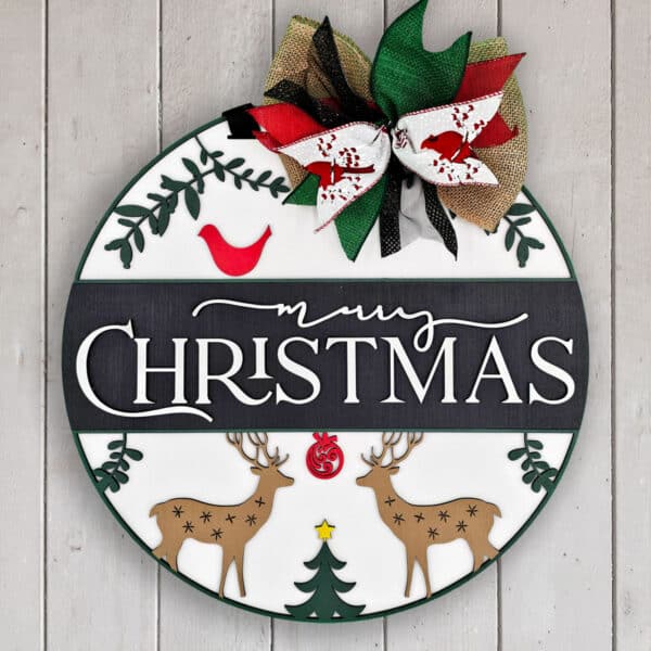 A round Christmas sign on a white wooden background features "Merry Christmas" in large white letters on a black strip. The sign is decorated with a red bird, two scenic deer, a Christmas tree, and topped with a festive bow in red, green, black, and burlap.