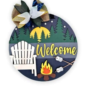 A circular welcome camper sign with a camping theme featuring trees, stars, a tent, an Adirondack chair, a campfire with marshmallows on sticks, and a large decorative bow at the top. The word "Welcome" is prominently displayed in yellow, making it the perfect wood door sign for nature lovers.