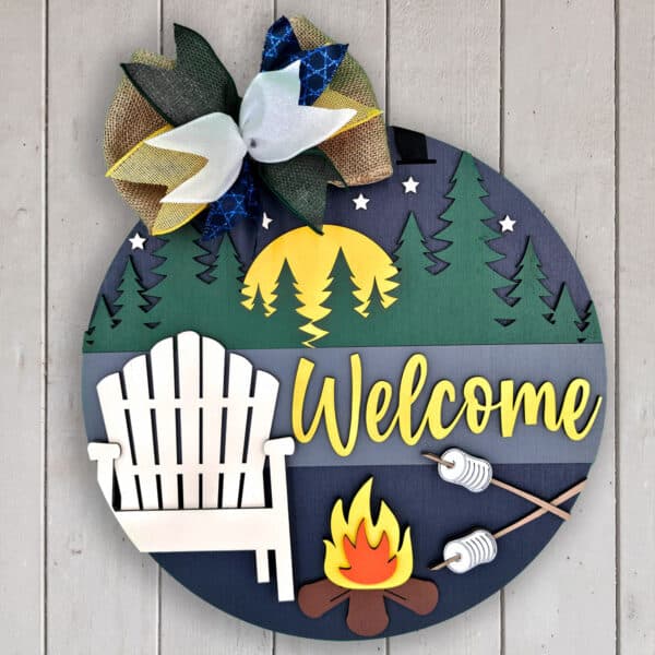 A round wooden door sign with a forest and night sky design features a large white bow at the top. Below, a white Adirondack chair sits next to a campfire with marshmallows on sticks, and the word "Welcome" is written in yellow.