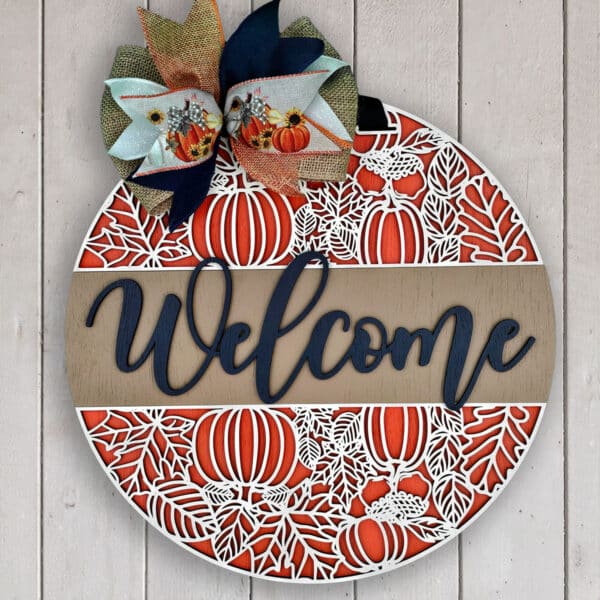 A round wooden custom door sign with intricate pumpkin and foliage designs in orange and white. The top has a large decorative bow featuring pumpkins and a sunflower print. The word "Welcome" is elegantly written across the center in bold, dark blue letters.