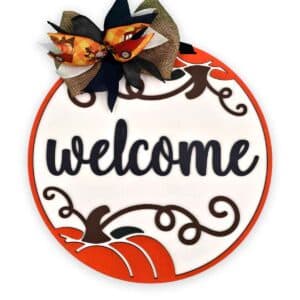 A round door sign with an orange border featuring the word "welcome" in large, black cursive letters. Decorative swirls and a pumpkin shape adorn the lower section, with a large fall-themed bow at the top in black, orange, and burlap colors. This welcome pumpkin round is perfect for autumn decor.