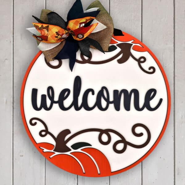 A round wooden door sign with an orange border and a pumpkin design at the bottom. The word "welcome" is written in large, black cursive letters in the center. At the top, there is a decorative bow made of various fabrics with an autumn theme. The background is a white wooden panel.