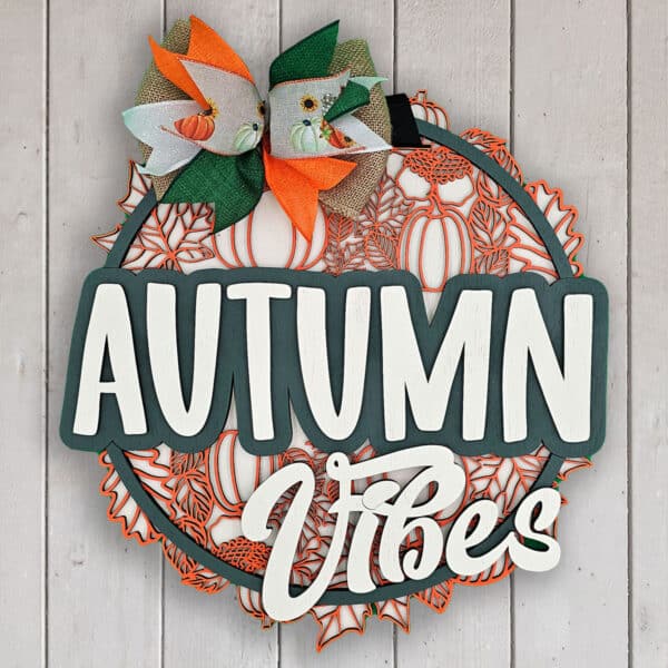 A decorative sign on a wooden background reads "Autumn Vibes" in white letters. It features a green frame adorned with a multicolored bow and pumpkin designs, embodying fall-themed colors of orange, green, and brown.