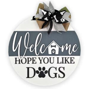 This charming round wooden door sign warmly greets visitors with the text, "Welcome Hope You Like Dogs." It showcases a small doghouse and playful paw prints, beautifully embellished with a burlap bow adorned with paw print ribbons.