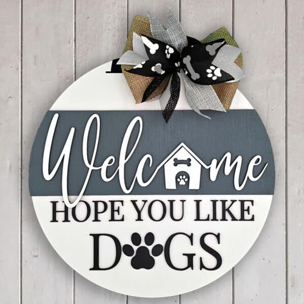 A round wooden welcome sign hangs against a white wall. The text, "Welcome, Hope You Like Dogs," is accented with paw prints and a dog house image. A patterned ribbon bow at the top adds charm, adorned with even more paw prints.