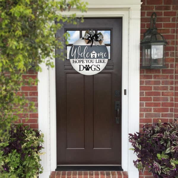 A dark brown front door with a glass panel at the top, set in a brick wall, surrounded by white trim. Above the door, a custom door sign reads, "Welcome, Hope You Like Dogs," with dog paw prints and a small house illustration. A garden lantern is on the right wall.